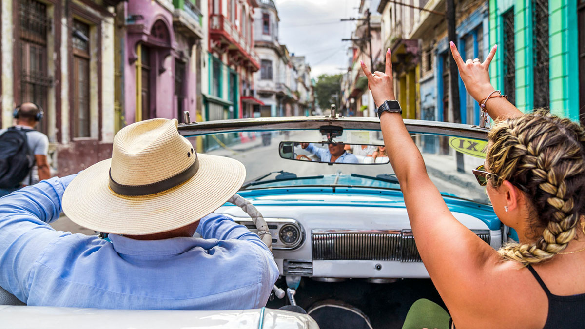 Man driving a blue car along the streets with a blonde woman in the passenger seat.