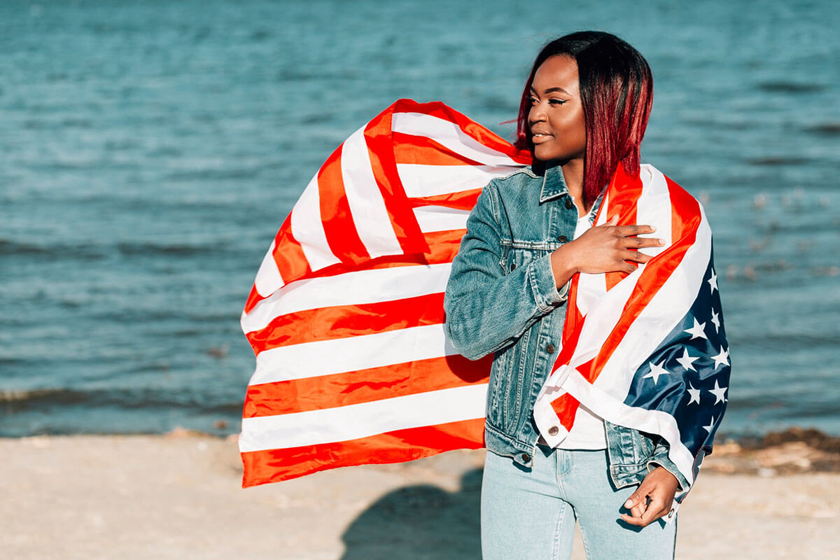 An American woman standing by the seashore with the American flag placed over her left shoulder and left arm.